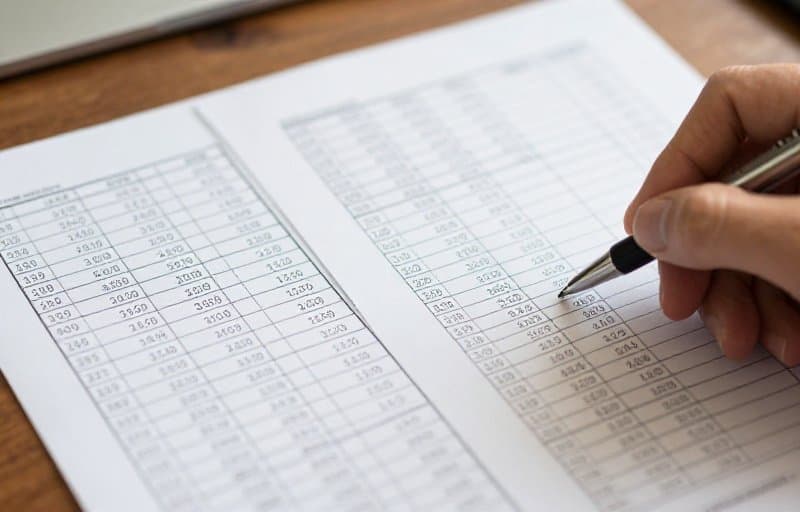 Two printed spreadsheets laid side by side while a hand traces one row across both, illustrating reconciling refund reports by transaction id