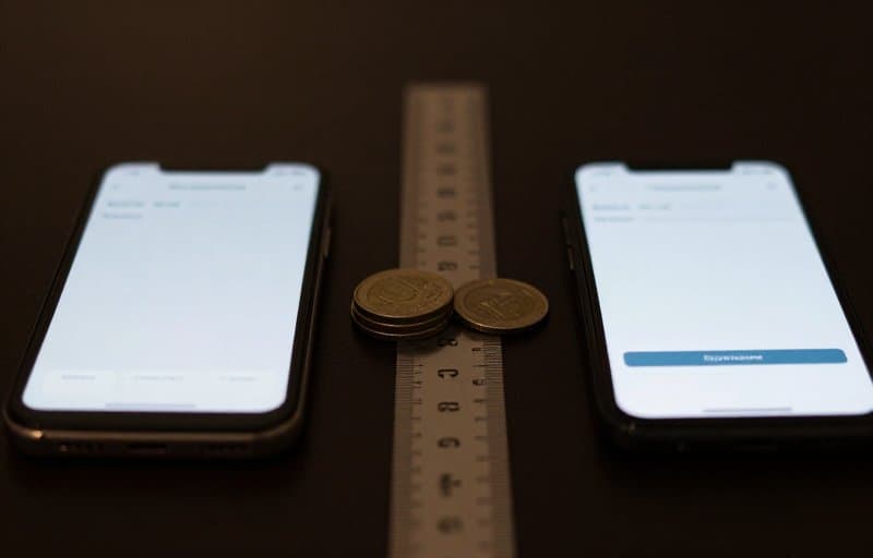 Dua ponsel pintar berdampingan di atas meja gelap menampilkan layar pembayaran langganan dengan setumpuk koin yang dikembalikan sebagian di antaranya, menggambarkan pengembalian dana langganan penuh versus prorata di App Store dan Google Play