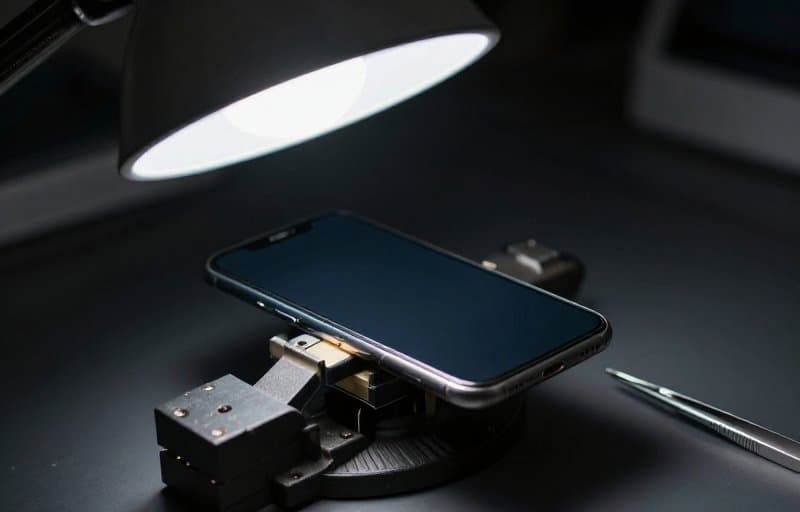 Sebuah ponsel pintar dijepit di ragum bangku kecil di bawah lampu kerja dengan pinset di sampingnya, sebuah perangkat yang sedang diuji mewakili latihan refund sebelum menjadi nyata